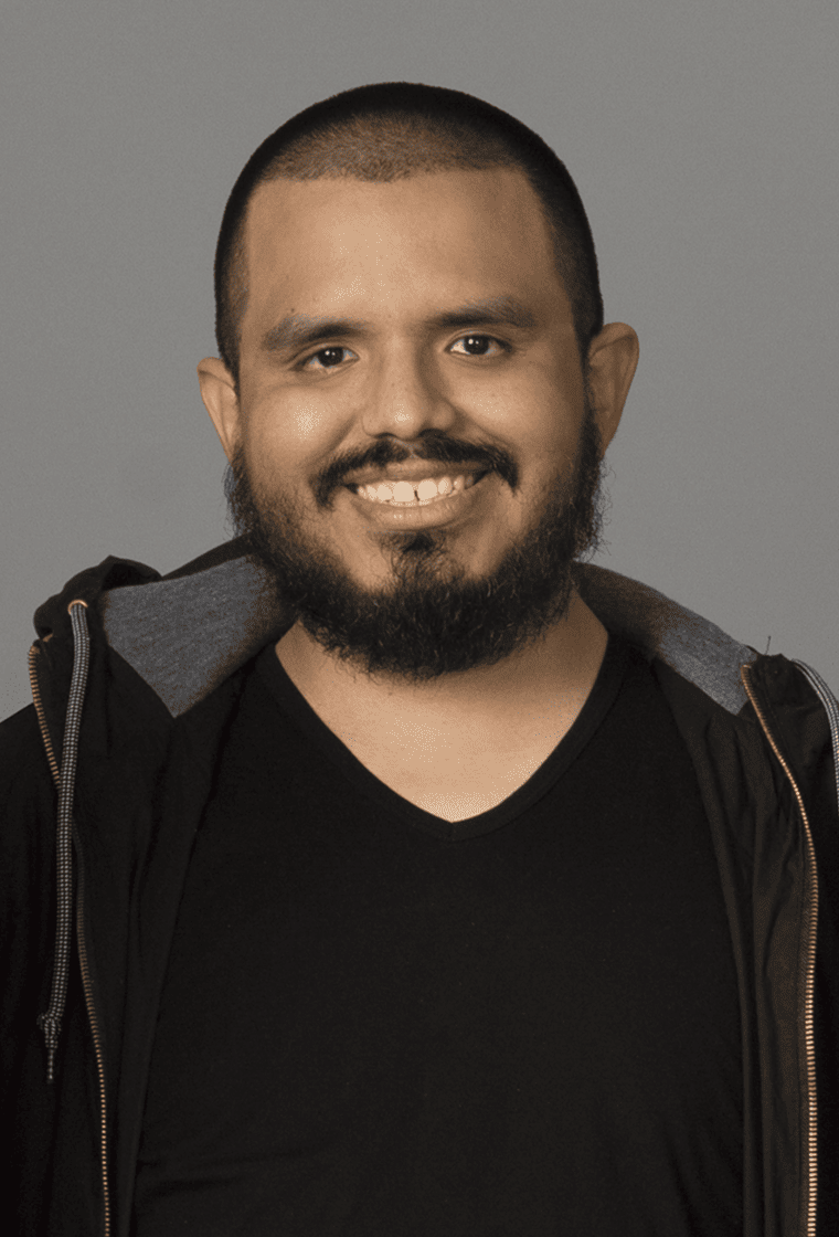 A man with a beard and short hair is smiling and wearing a black shirt and a black jacket with a gray hood.