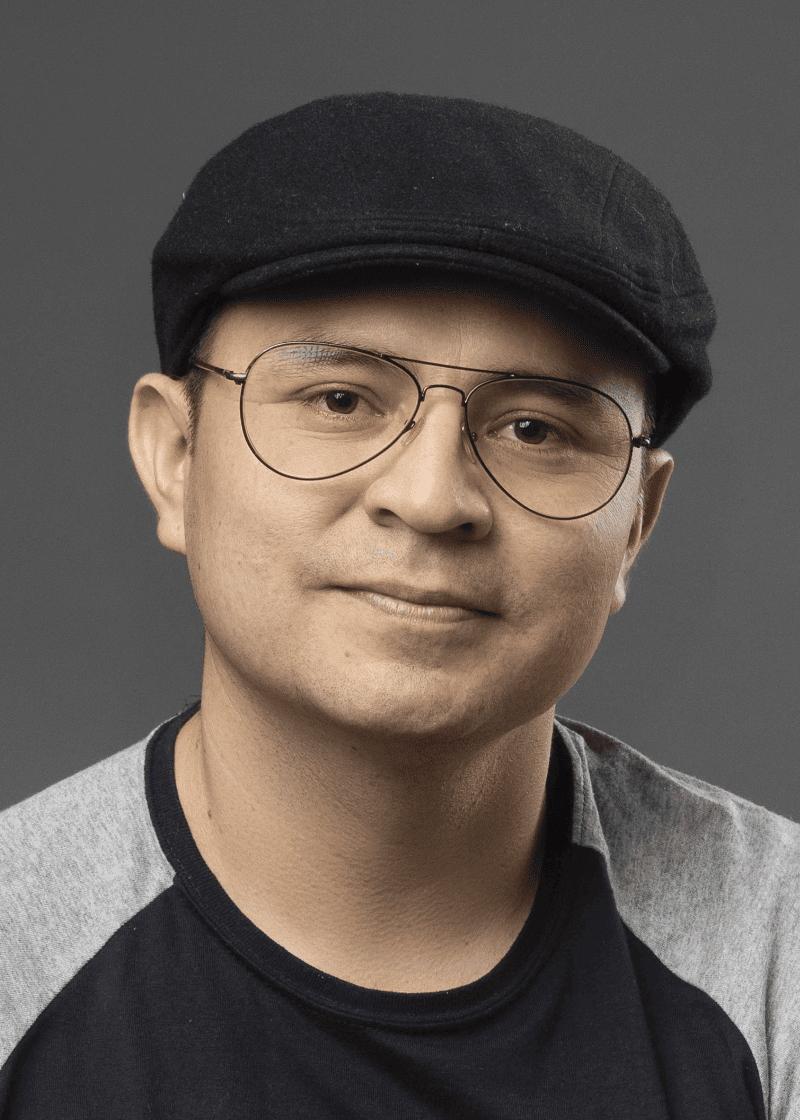 Alex Aliaga A man wearing a black cap, round glasses, and a black and gray shirt smiles softly against a gray background.