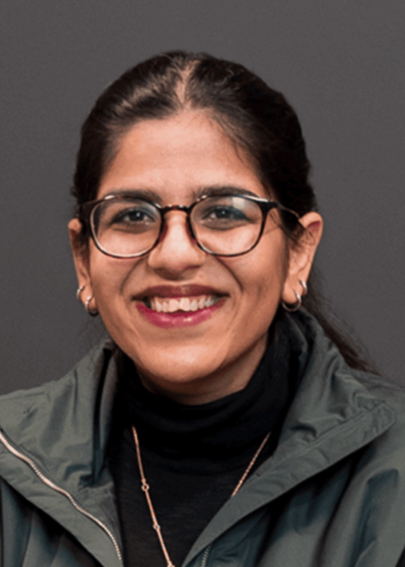 Avval Kaur A woman with glasses, earrings, and a black turtleneck smiling against a gray background.