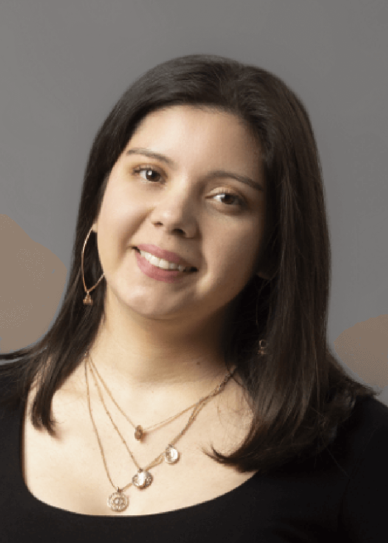 Carmela Cabrera A woman with shoulder-length dark hair, wearing a black top and layered gold necklaces, smiles at the camera against a gray
