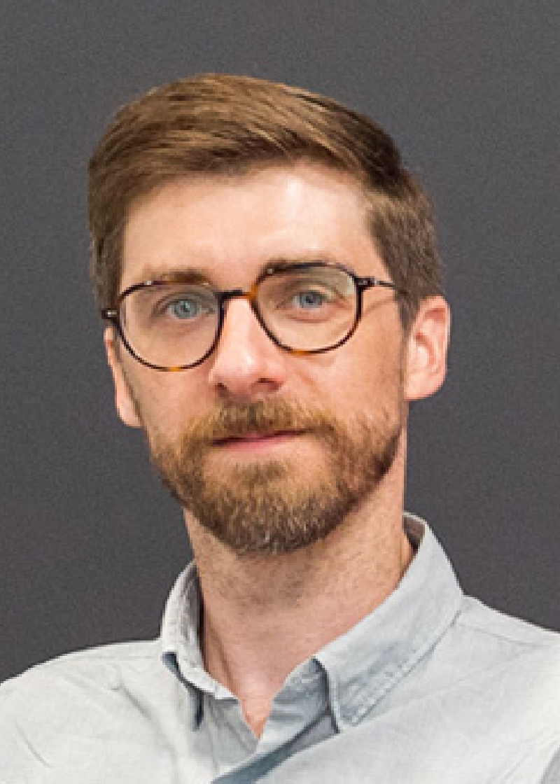 Danny McGarvey A man with brown hair, glasses, and a beard wearing a light gray shirt against a dark background.