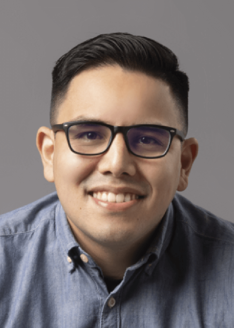 Jean Pierre Ruiz A smiling man with short dark hair, glasses, and a denim shirt poses against a gray background.
