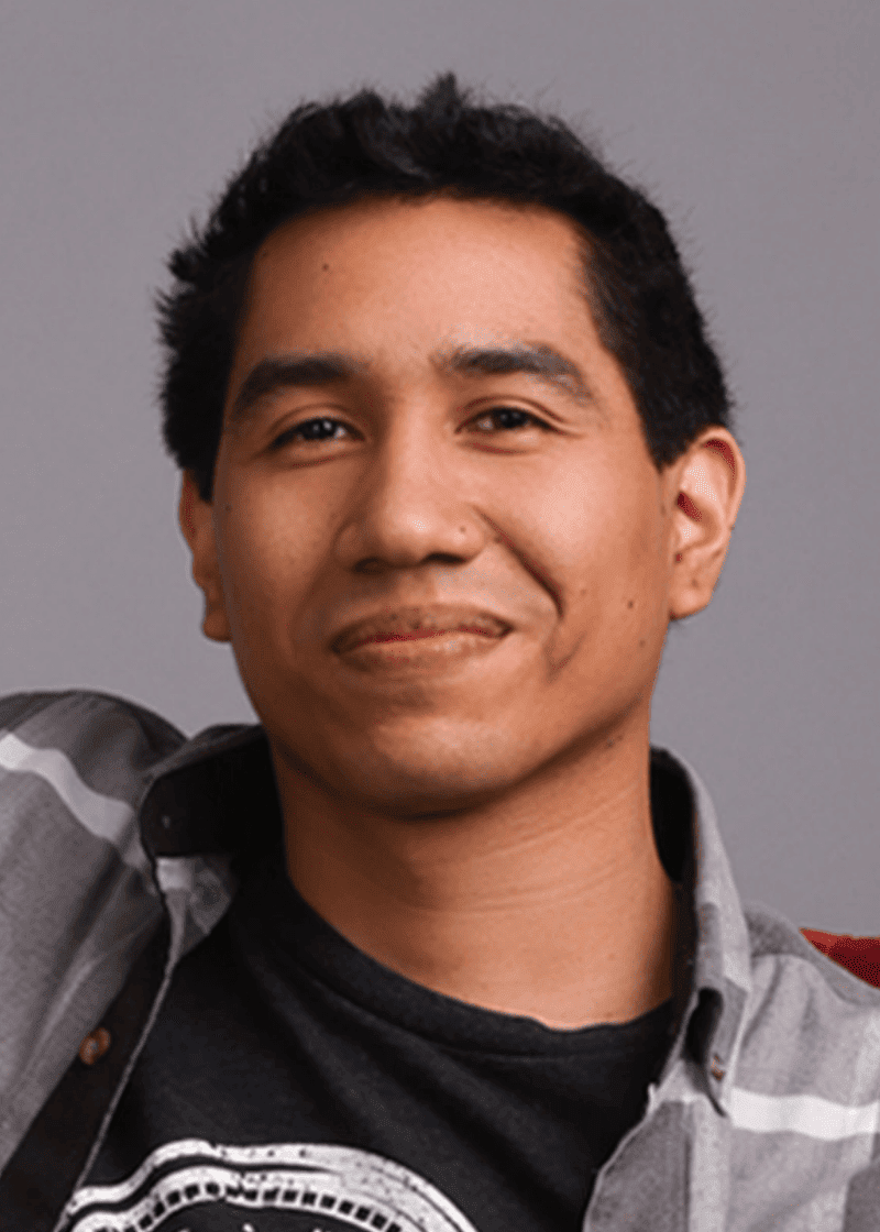 Manuel Francia A young man with short black hair, smiling slightly, wearing a black t-shirt and a gray plaid shirt, against a gray