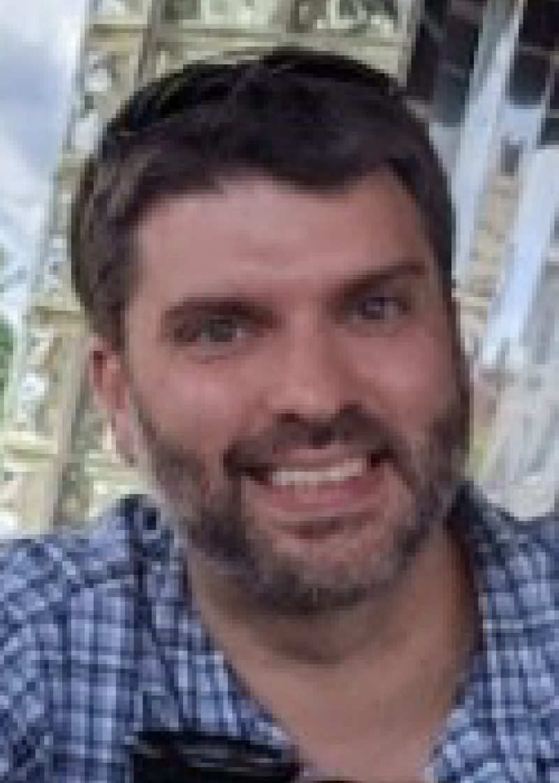 Sam Salyards A smiling man with dark hair and a beard, wearing a blue checkered shirt, outdoors near a metal structure.
