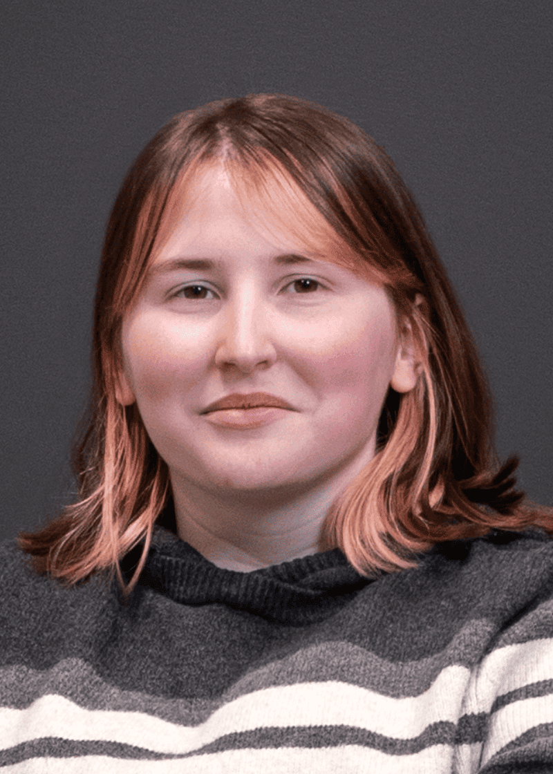Shelby Baker A woman with shoulder-length reddish-brown hair and a subtle smile, wearing a black and white striped sweater, poses against