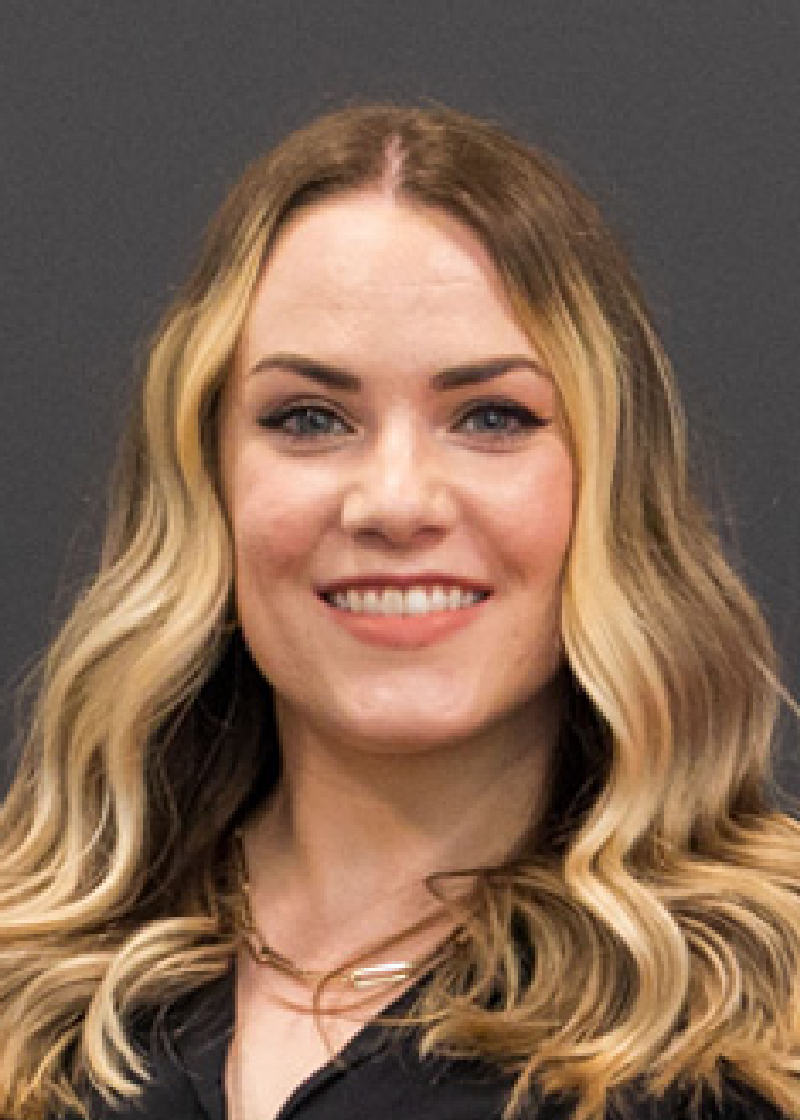 Sydney Meyer A woman with long, wavy blonde hair, blue eyes, and a bright smile wearing a black top and layered necklaces.