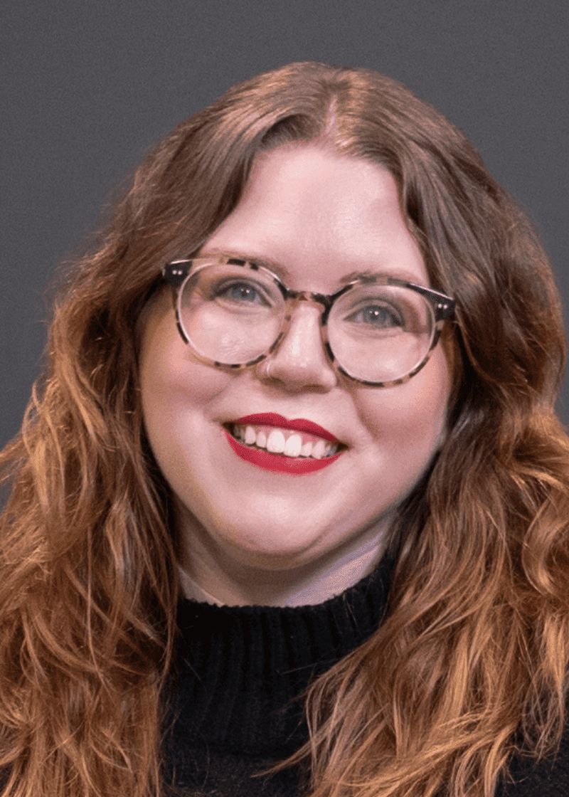 Tyler Drummond A woman with long, curly red hair, wearing glasses and red lipstick, smiles against a dark background.