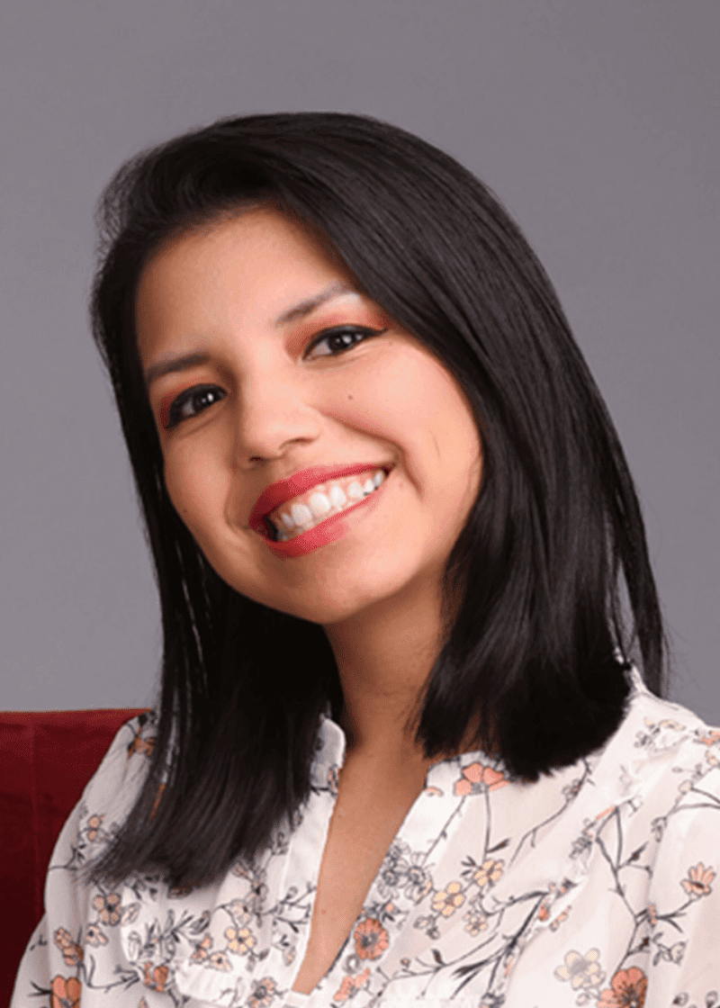 Vannia Ochoa A woman with shoulder-length black hair, wearing a floral blouse, smiling against a gray background.