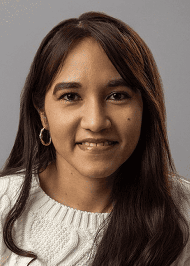 Ivonne Francia A woman with long, dark brown hair, wearing a white knitted sweater and gold hoop earrings, smiles at the camera against a