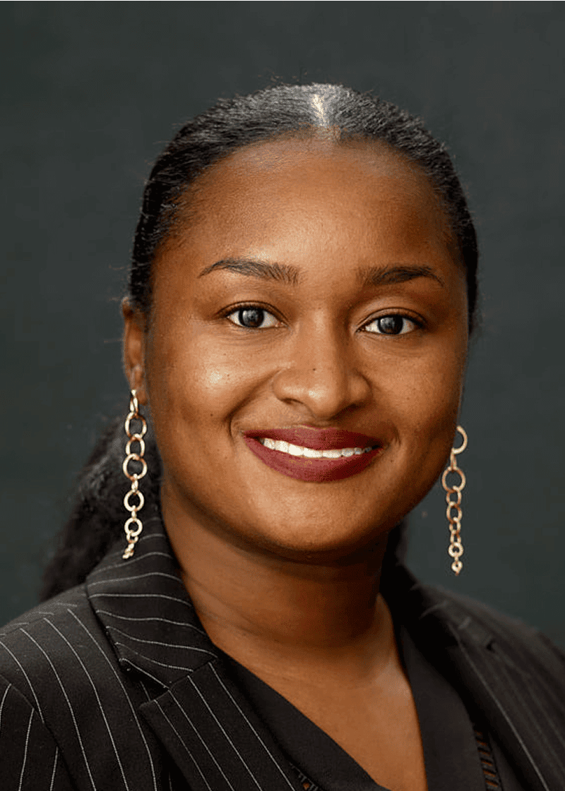 Domanique Cummings A woman with dark hair in a ponytail, wearing a pinstripe blazer and gold dangling earrings, smiling against a dark