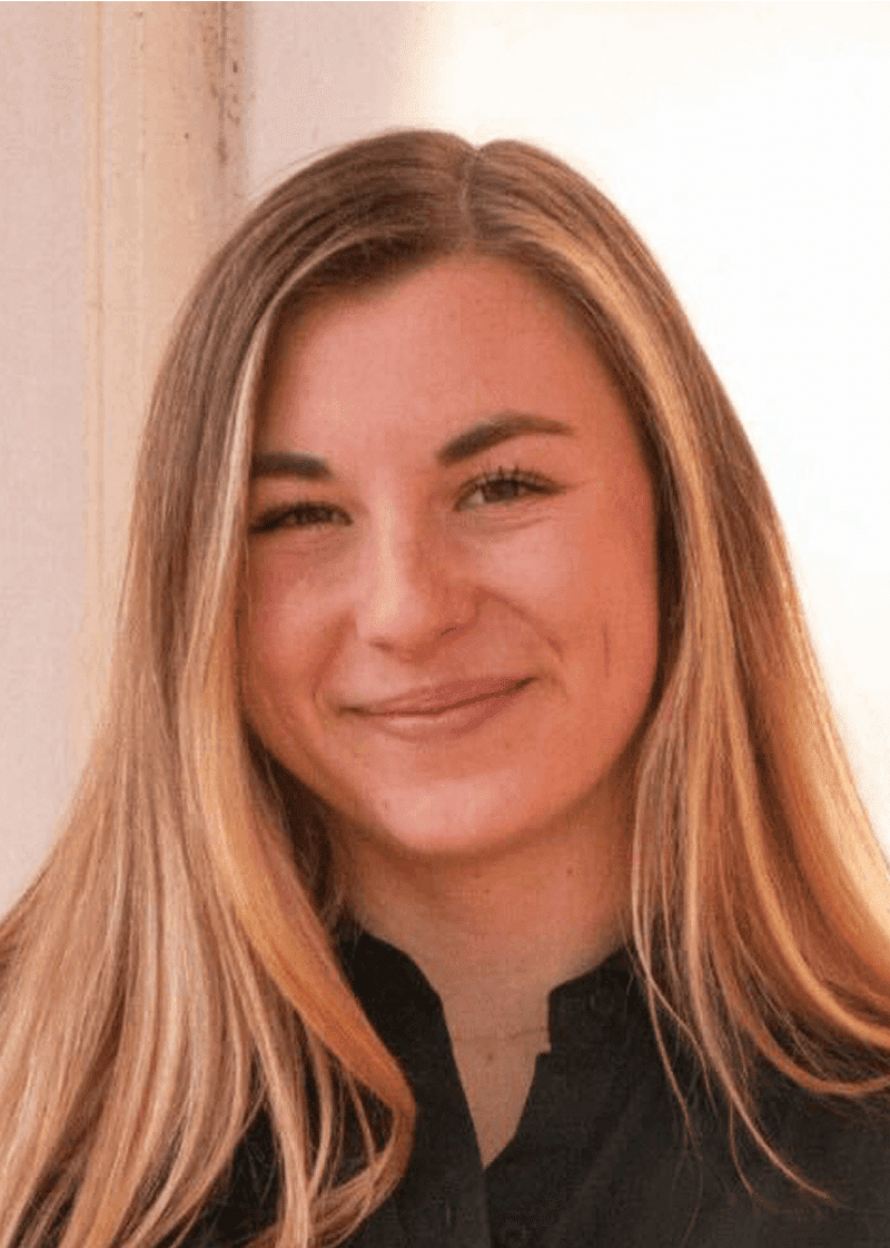 Sydney Jones A woman with long, wavy blonde hair, smiling softly, wearing a black top, standing near a light-colored wall.