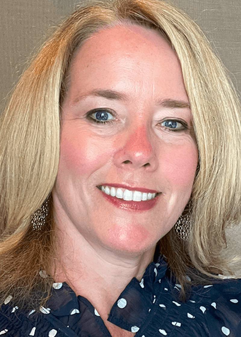 Caron Beesley A woman with blonde hair, blue eyes, and a bright smile, wearing a dark polka-dot blouse and earrings.