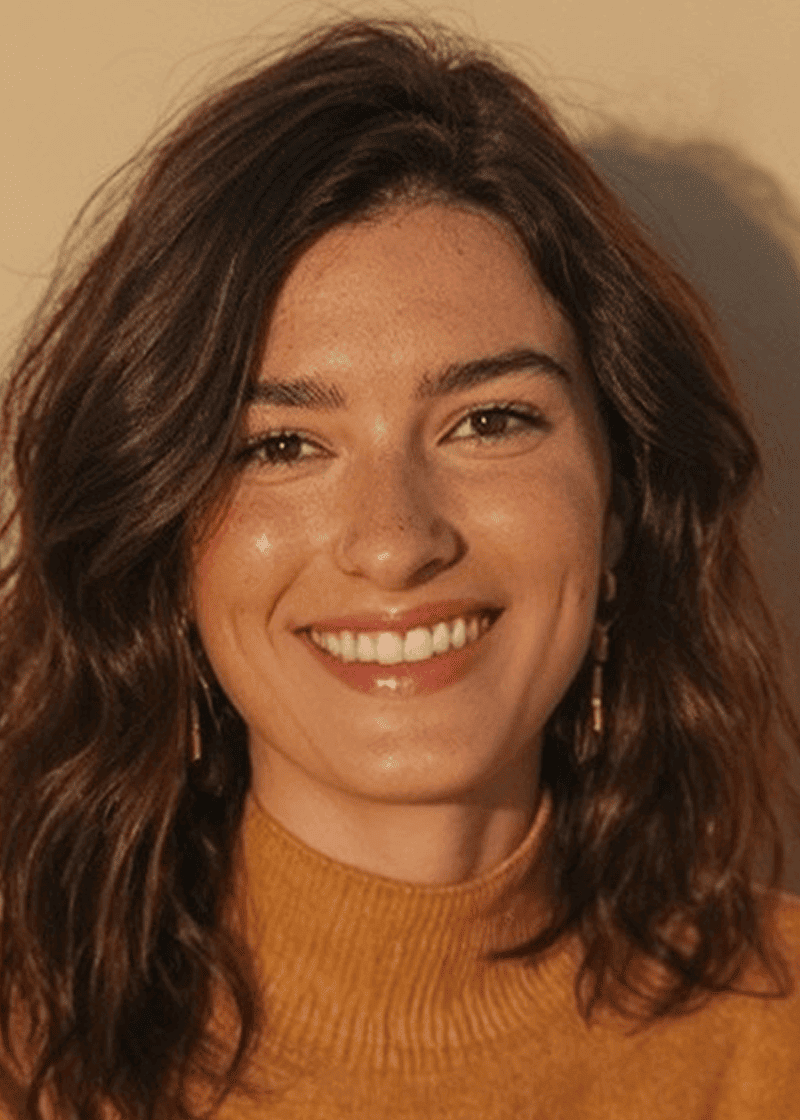 Makenzie Smith A woman with wavy brown hair, wearing a mustard turtleneck and earrings, smiling warmly at the camera.