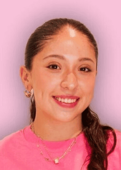 Karen Buenrostro A young woman with long brown hair in a ponytail, smiling, wearing a pink top, earrings, and a gold necklace against a pink