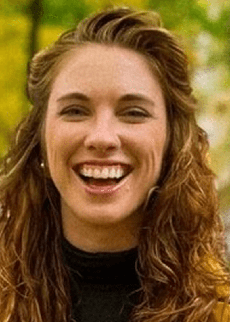 Kira Johnston A woman with curly, reddish-brown hair smiling outdoors with a blurred green background.