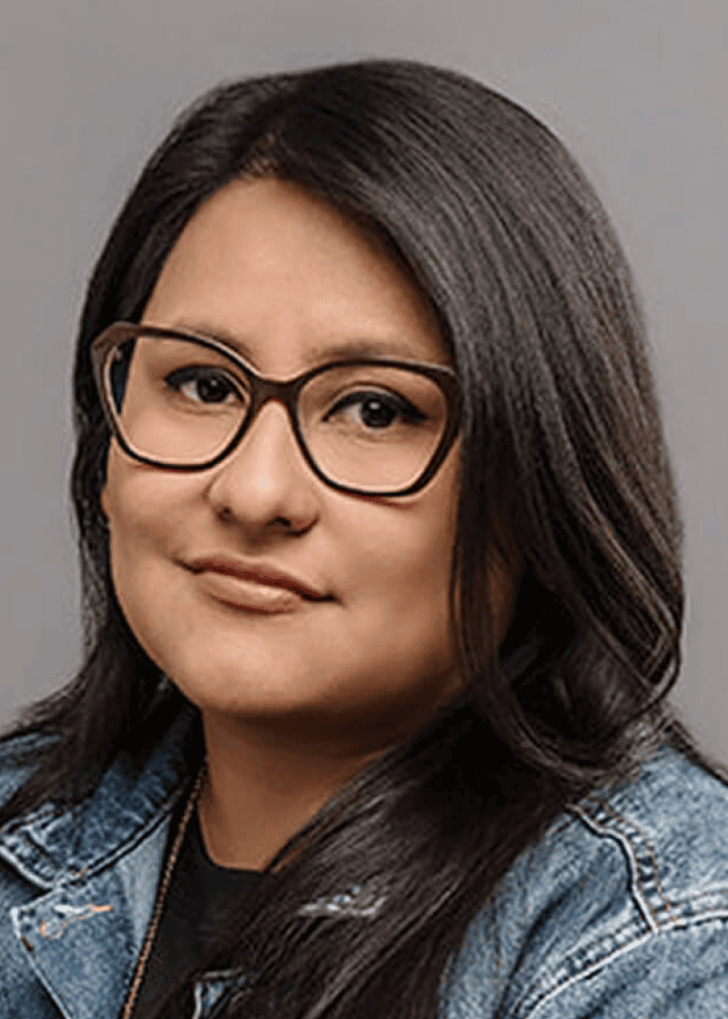 Brenda Mendoza Woman with long dark hair, glasses, and a denim jacket, smiling softly against a gray background.