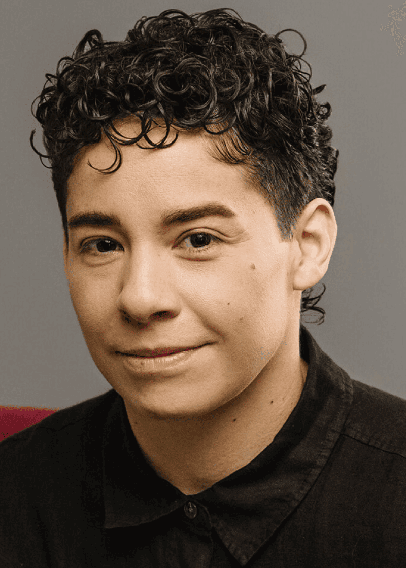 Janine Bosworth A person with short, dark, curly hair, light skin, and a subtle smile, wearing a black collared shirt against a gray
