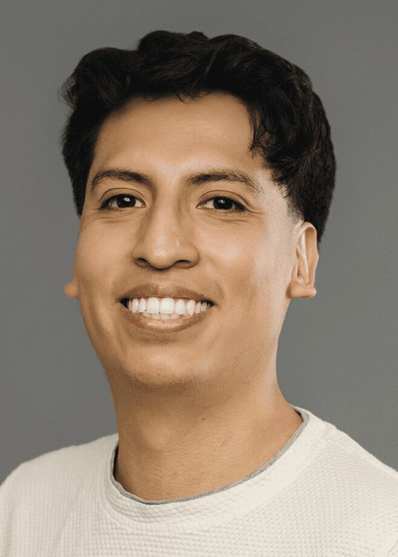 Ulises Oliveros A young man with dark, wavy hair smiling against a gray background, wearing a light-colored textured shirt.